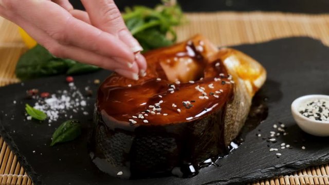 Salmon steak teriyaki with sesame seeds on black slate background. Asian food cooking, female chef preparing fish for grilling. Slow motion of seasoning fish