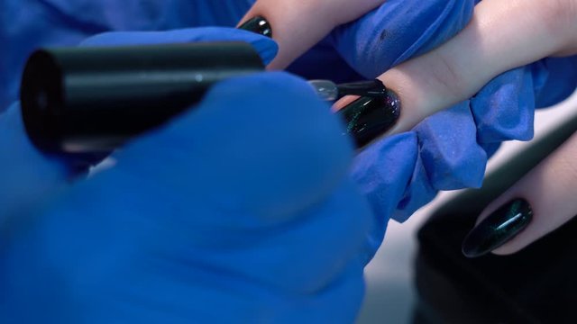 Closeup view of beautiful manicured nails of woman and professional manicurist applying transparent finish top polish laer of gel polish. Black design with shiny sparkling cat eye effect. 4k video.