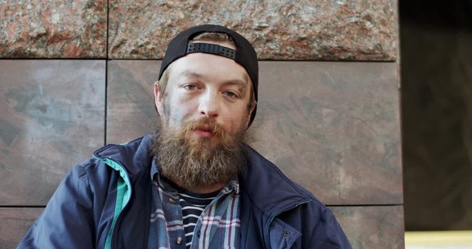 Portrait of the young Caucasian homeless man in hat and poor ugly look looking straight to the camera with tired eyes. Outdoors.