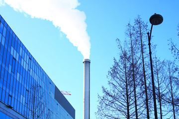 Close-up on an industrial chimney. Polluting smoke in urban areas. Clear sky in the background. Building and vegetation without foliage in the foreground.