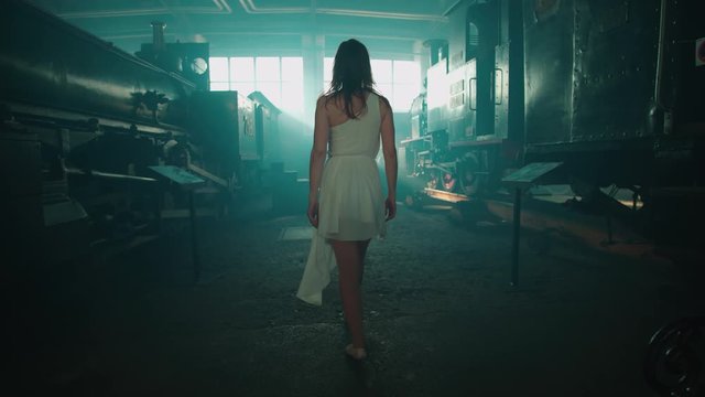 Following woman between trains in smoke 4K. Wide shot gimbal stabilizer walking behind a person walking away in focus inside the hall with locomotives. Bright background sun flares.