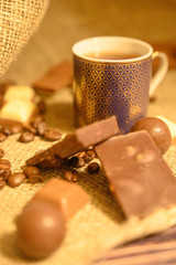 Chocolate candies, coffee beans and a mug of tea on a background of rough homespun fabric. Close up. Vertical position.