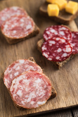 Sandwiches with smoked salchichon and braunschweig sausages are on wooden board. Next to are cheese cubes. Close up, vertical composition.