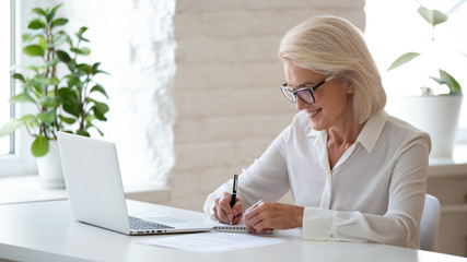 Mature businesswoman sitting at desk makes notes writing on notepad - Powered by Adobe