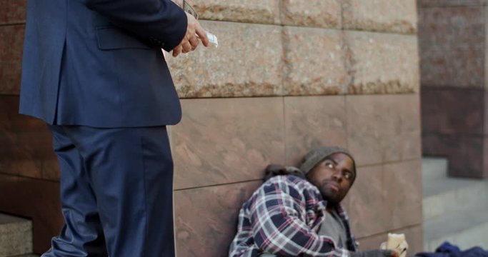 Caucasian rich businessman walking by the African American homeless man and giving him 100 dollars banknote, so beggar is very happy and surprised.