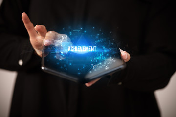Businessman holding a foldable smartphone with ACHIEVEMENT inscription, business concept