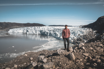 Fototapeta premium A young man traveler tourist standing in front of Eqip Sermia glacier called Eqi Glacier. Wall of ice in background. The concept of global warming and professional guides. On a sunny day with blue sky