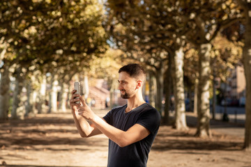 Young man in tshirt standing on street and makes photo on mobile phone. In the background is a tree lane. Frankfurt, Germany