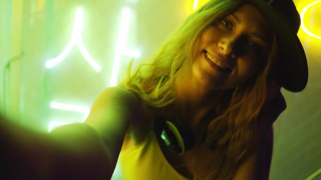 Hip-hop girl in cap posing against colored neon lights. POV portrait of teenage woman in cap looking at camera while taking selfie in dark interior of nightclub. Nightlife, disco, concept
