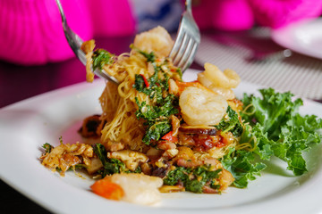 Close up view of using spoon and fork catch Thai style of Stir fry Spicy basil with rice noodle, vermicelli, and shrimp, on white dish.  