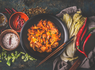 Homemade kimchi in black bowl on rustic background with ingredients: chinese cabbage , chili, spices and salt . Top view. Healthy fermented food - obrazy, fototapety, plakaty