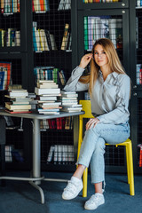 Pretty blonde woman or model  sitting in college library with books on table, holding glasses.