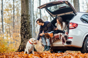 Young couple have a picnic with their dog near automobile in the autumn forest.
