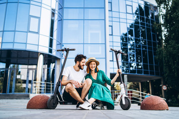 Fototapeta premium Young loving couple relaxing sitting near a modern glass building with their electric scooters. Walks in the big city