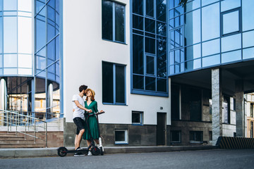 A young smiling couple enjoy a walk in the city on electric scooters. Content technologies. Large modern building on background