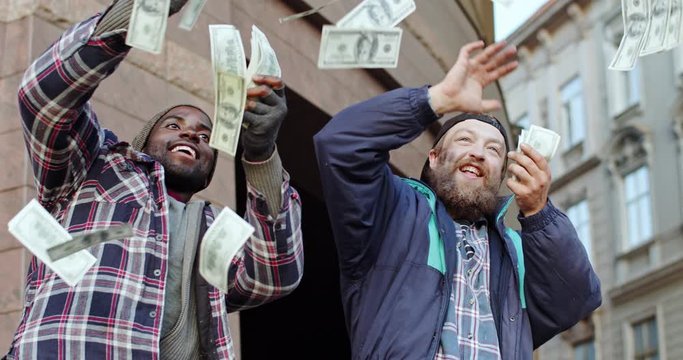 Two mixed-races happy cheerful homeless men laughing and throwing dollars banknotes as now hace a lot of money and becoming rich.