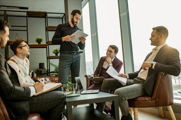 young caucasian bearded man in black formal shirt stand giving speech to co-workers, holding paper document in hands. isolated in modern office with panoramic window