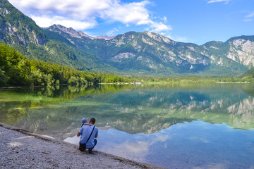 Father and baby admire calm mountain lake on a bright sunny day. Man hugs son on a lake birch, and looks into the distance.