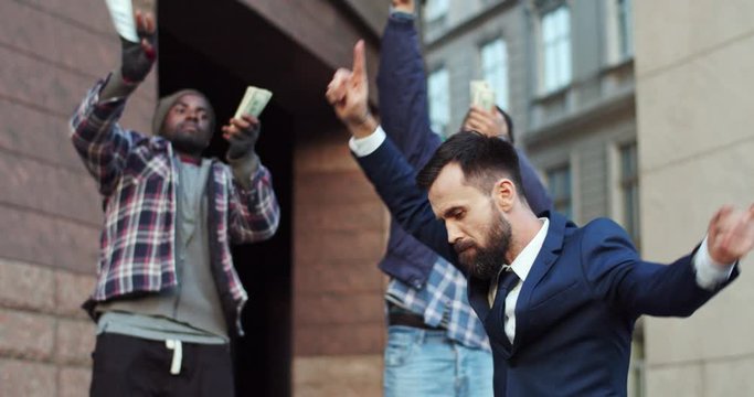 Multiethnic three young men dancing and having fun together at the street as throwing money in the air, two homeless guys and rich businessman are friends. Tossing banknotes in the air.