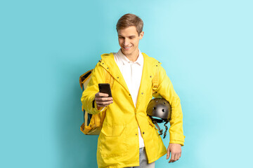 portrait of young smiling caucasian man working in express delivery service, driving motorcycle, holding helmet and wearing yellow uniform look at smartphone isolated over blue background