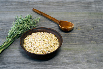 Oatmeal flakes in a ceramic brown bowl with a wooden spoon and ears of oats on a rustic wooden table background.