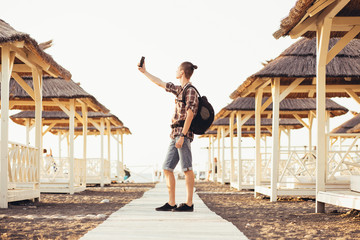 Young guy tourist in casual clothes with a backpack makes selfie on the seashore on a sunny summer day. Travel concept and memory. Copyspace