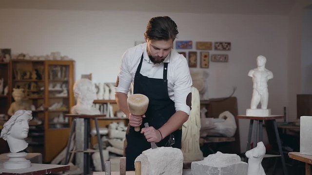 Handsome concentrated bearded sculptor beating on white limestone with hammer and chisel in his workshop
