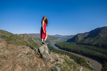 Naklejka premium Portrait of a woman in a red dress on top of a mountain on the background of the river flowing between the rocks.
