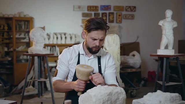 Attantive attractive bearded master sitting in his workshop and knocking off the limestone the necessary shape using chisel and mallet