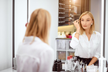 beautiful caucasian blond beautician in white shirt sit looking at mirror, at her work place in beauty salon.