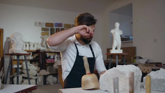 Slow motion of handsome professional young sculptor which having a rest while working in the craft studio with limestone and looking away
