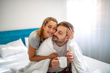 Obraz na płótnie Canvas Caring wife. Young woman taking care of his sick husband, bringing tea to bed. Couple with cold in bed at home together. Nice caring woman touching her husbands forehead while sitting near him