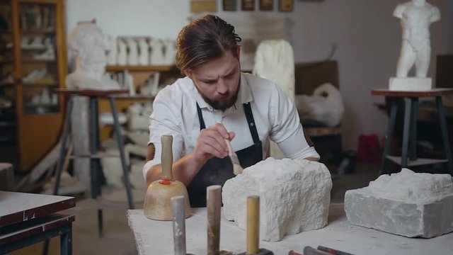 Attractive portrait of young talanted handsome sculptor which cleaning down the remains of limestone with brush on the craft studio background