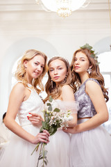 three young caucasian women with beautiful makeup and hair try on white wedding dresses together, dream to be bride, holding bouquet of flowers in hands and posing in salon among dresses