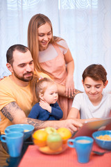 Obraz na płótnie Canvas love and family concept. kids and parents in casual wear in modern kitchen with modern interior, have meal and watching video on modern laptop