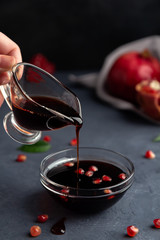 A hand pours pomegranate sauce from a gravy boat into a transparent bowl on a dark background