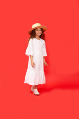 Cute little curly girl fashion pretty model with curly hair in straw hat and long white dress, looking at camera and laugh isolated red background.