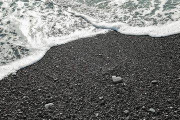 A gray stone heart-shaped lies on the shore with sea foam and clear water on a black sand beach. Concept of love, wedding, freedom. Background to Valentines day card with copy space.