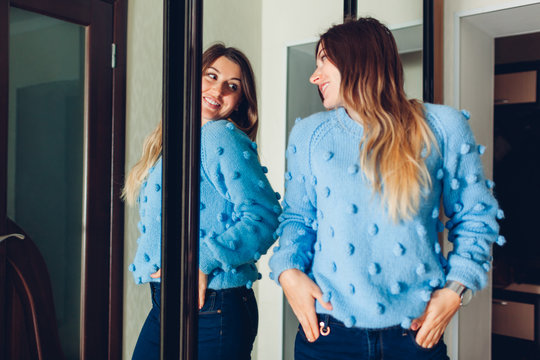 Woman in sweater looking at mirror. Girl trying clothes on by wardrobe at home. Beauty fashion