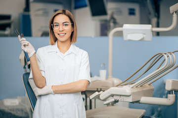 portrait of female dentist in white doctor's uniform standing in office near equipment and look at camera. Medicine, dentist, orthodontic concept