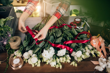 Fototapeta na wymiar top view on young woman hands tying flowers in red ribbon, preparing them for sale, wonderful white bouquet on table