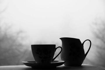 Black and white still life with cup, saucer and jug for milk on blurred background. Misty day in Ukraine. Loneliness concept. Copy space.