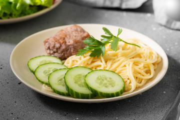 Pasta, cutlet and cucumbers on a beige plate on a gray table	