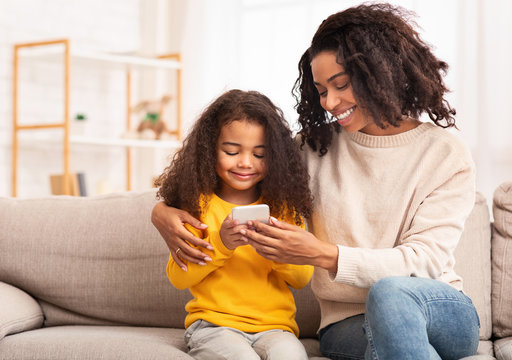 African Mother And Daughter Using Smartphone Sitting On Couch Indoor