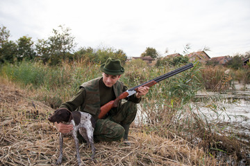 Hunter wearing green suit and hat holding with one hand his hunter dog