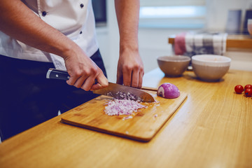 Close up of caucasian chef in uniform standing in domestic kitchen and chopping onion in chopping board.