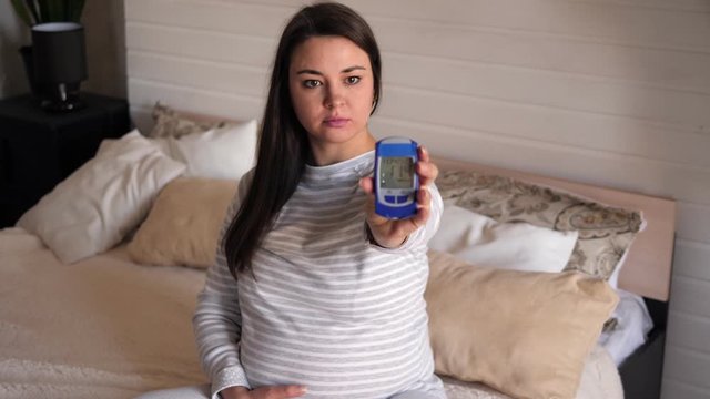Portrait of a pregnant woman with a blood glucose meter in her hands at home on the bed. A pregnant woman with diabetes measures her blood glucose level at home while sitting on a bed.