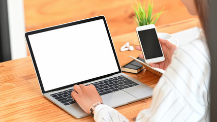 Behind shot of creative woman in stripped white shirt typing on white blank screen laptop and holding black blank screen smartphone in hand over wooden working desk background.