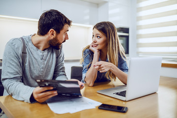 Young attractive bearded man holding electricity bill and showing to his girlfriend how much they spent. Woman pointing at bill and wondering.