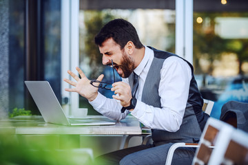 Obraz premium Angry elegant businessman in suit looking at laptop and yelling while sitting in cafe.